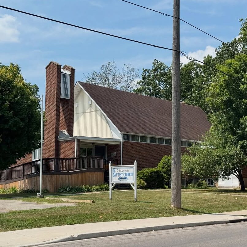 Red-brick Baptist church with steep roof, bell tower, and ramp amid trees.