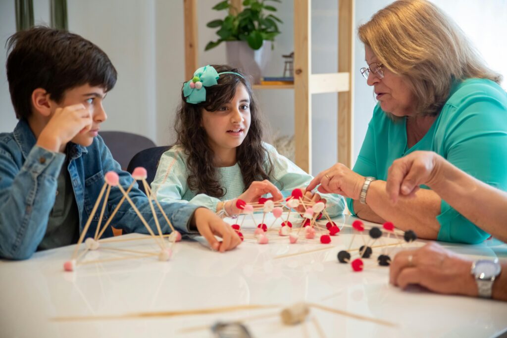 Children and an older woman build pom-pom and stick structures at a table.