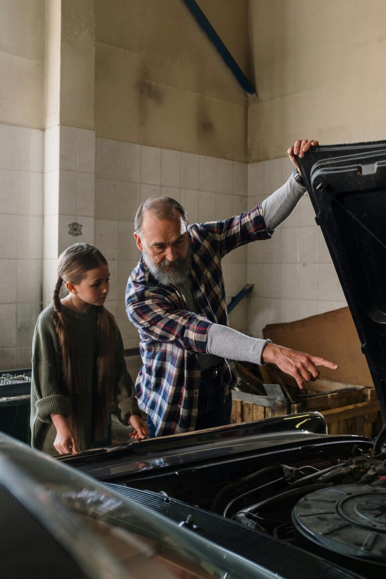 Older man points under a car hood as a girl looks on.