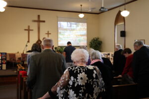 Congregation standing in pews during a church service, facing a stage with wooden crosses and a projected hymn on a screen.