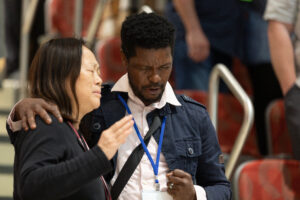 Two attendees with lanyards pray together, eyes closed, in a large indoor gathering.