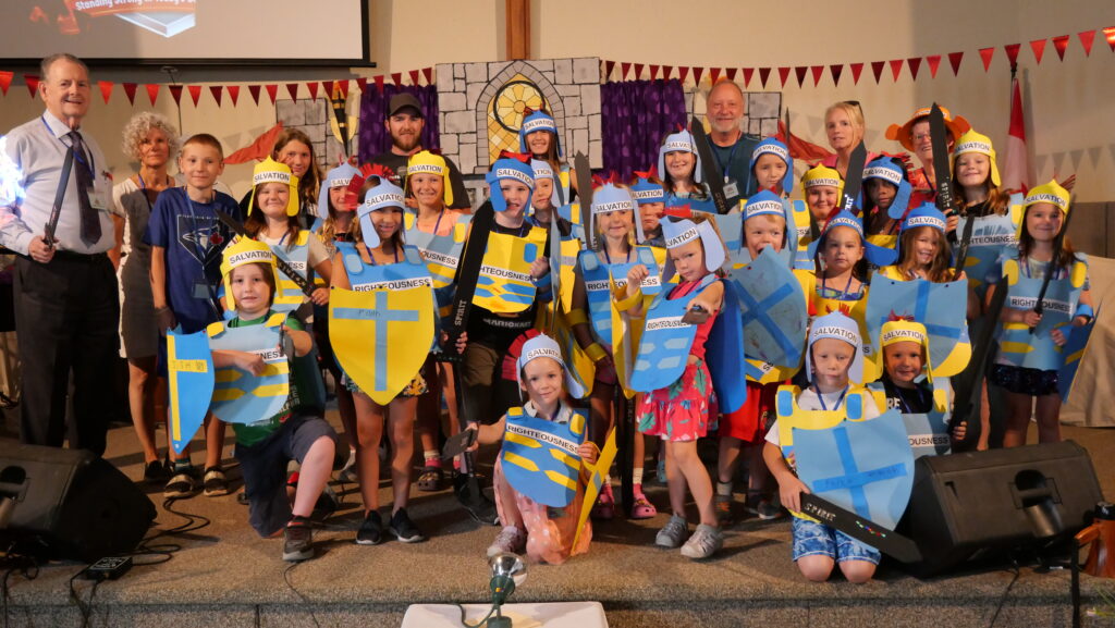 Group of children in paper knight costumes holding shields on a stage, with adult leaders behind them