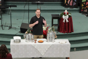 Speaker with microphone at church behind a communion table with trays, bread, cup, and Advent candles.