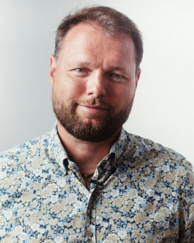 Headshot of a bearded person in a blue floral shirt with a lapel microphone against a light background