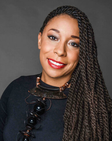 Smiling person with long braids in a black top and bold black-and-copper statement necklace on a gray background