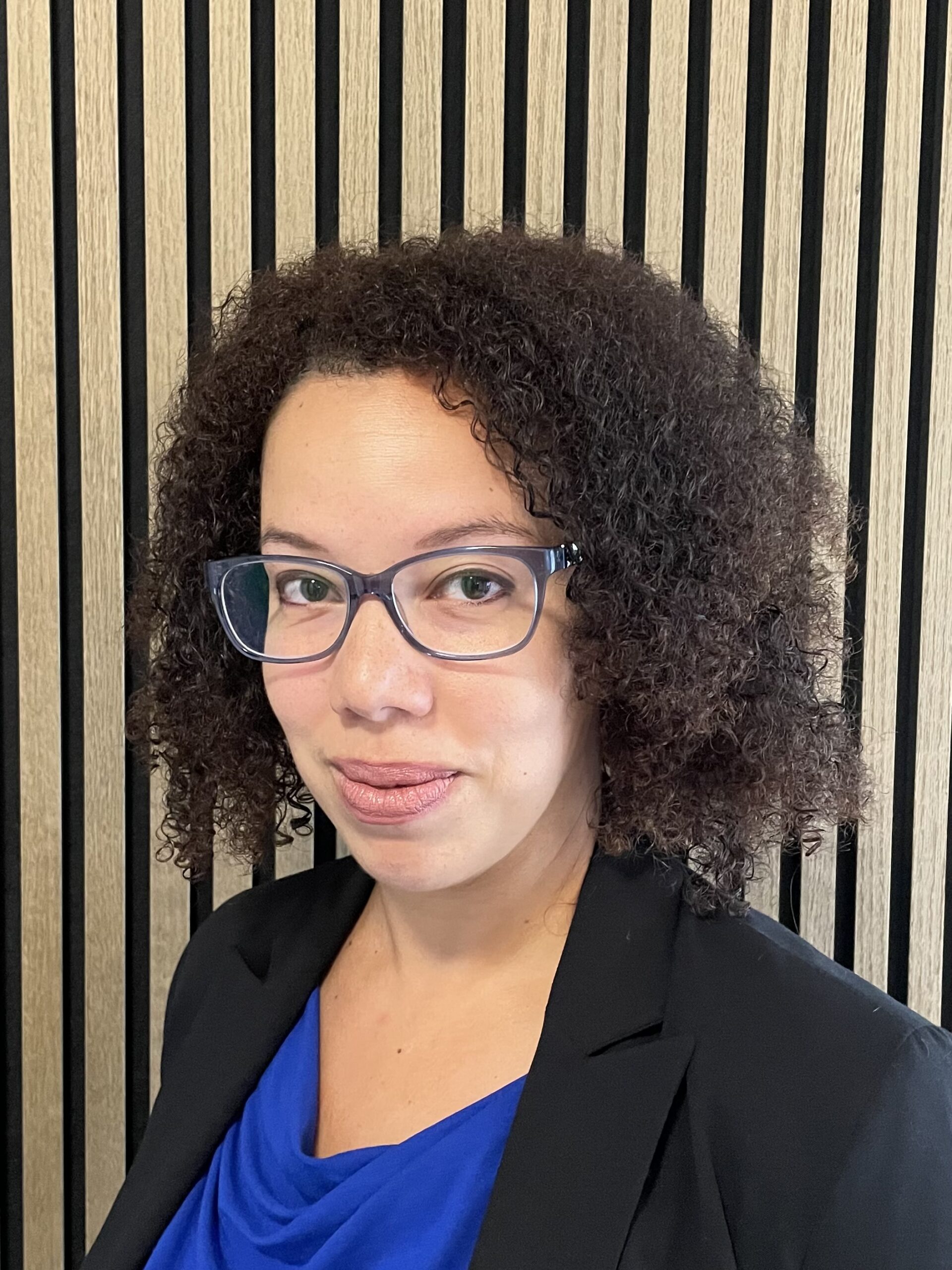 Professional headshot of a person with curly hair and glasses in a blue top and black blazer against a wood slat wall.