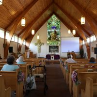 Wooden vaulted church with stained glass, flower-decorated pews, and a few seated people.