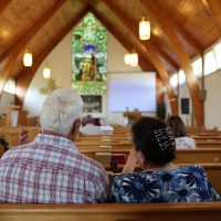Elderly couple seated in wooden church pews facing altar and stained glass.